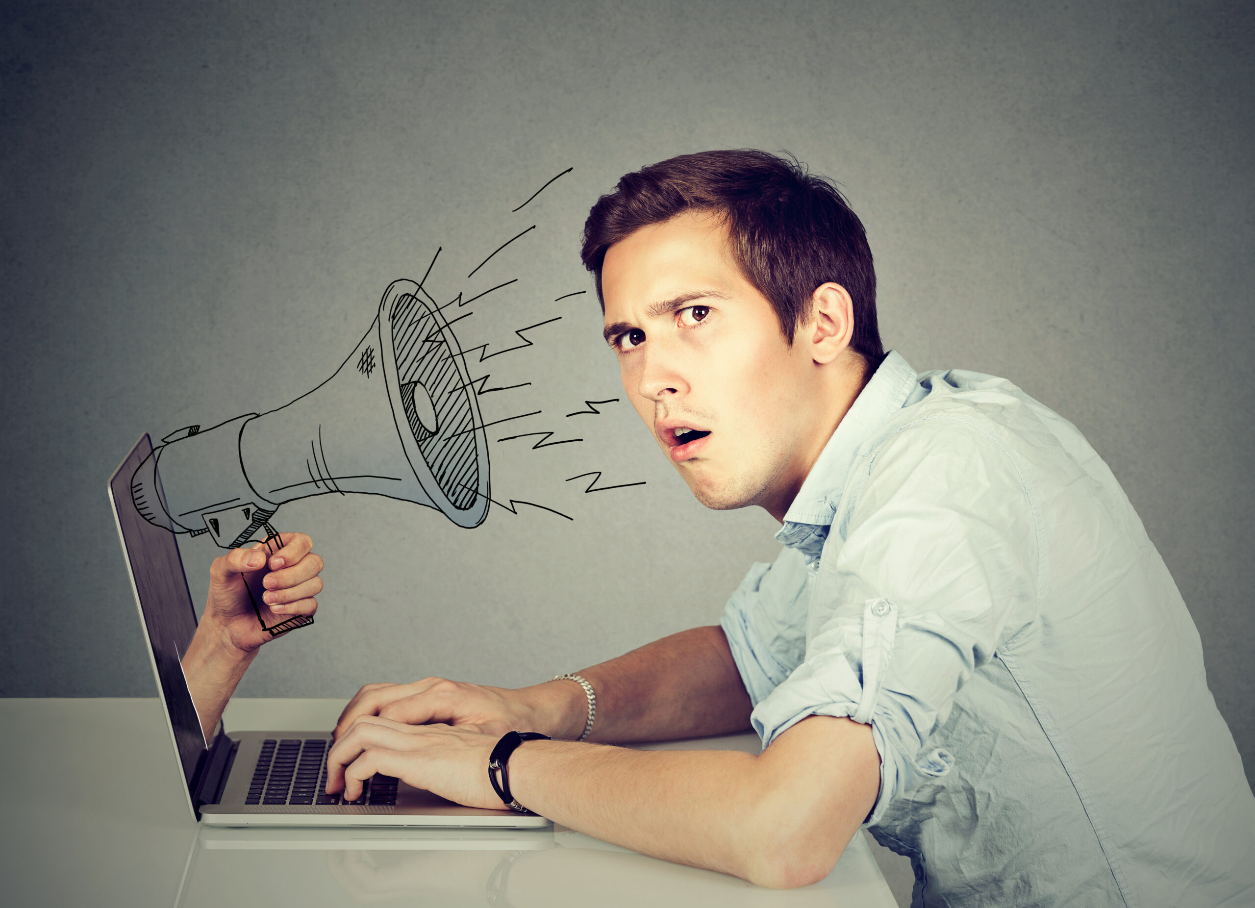 Side profile young anxious man sitting at table using working on a computer with megaphone poking out from a laptop screen